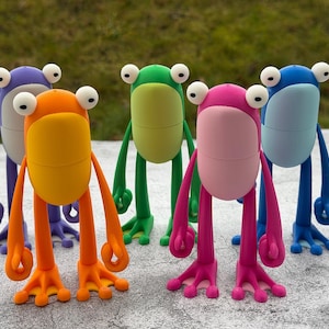 May include: A group of five colorful frog figurines. The frogs are in shades of orange, purple, green, pink, and blue. Each frog has large white eyes and long, thin limbs. The figurines are standing on a gray surface.