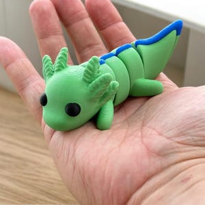 May include: A green and blue 3D printed axolotl figurine with black eyes. The figurine is posed with its head down and its tail curled up.