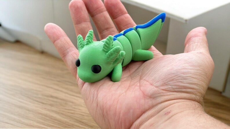 May include: A green and blue 3D printed axolotl figurine with black eyes. The figurine is posed with its head down and its tail curled up.