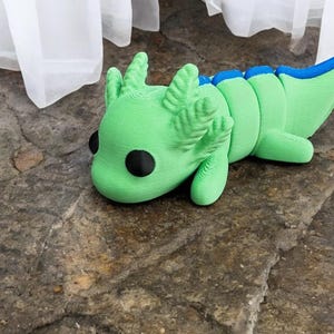May include: A green and blue axolotl figurine with large black eyes. The figurine is lying on its side with its head resting on the ground.