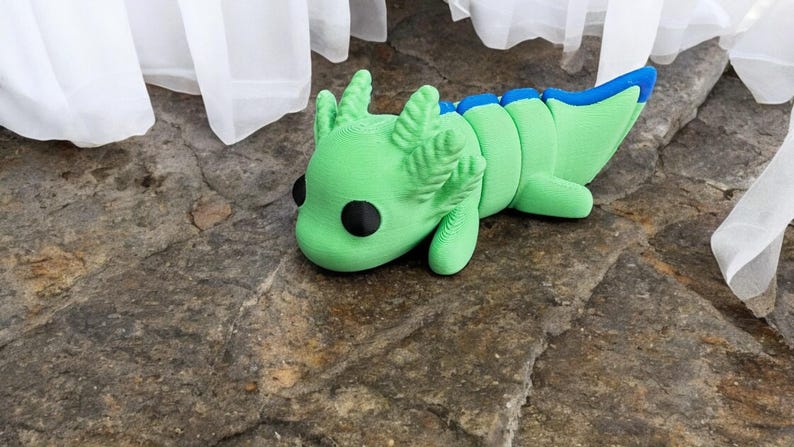 May include: A green and blue axolotl figurine with large black eyes. The figurine is lying on its side with its head resting on the ground.