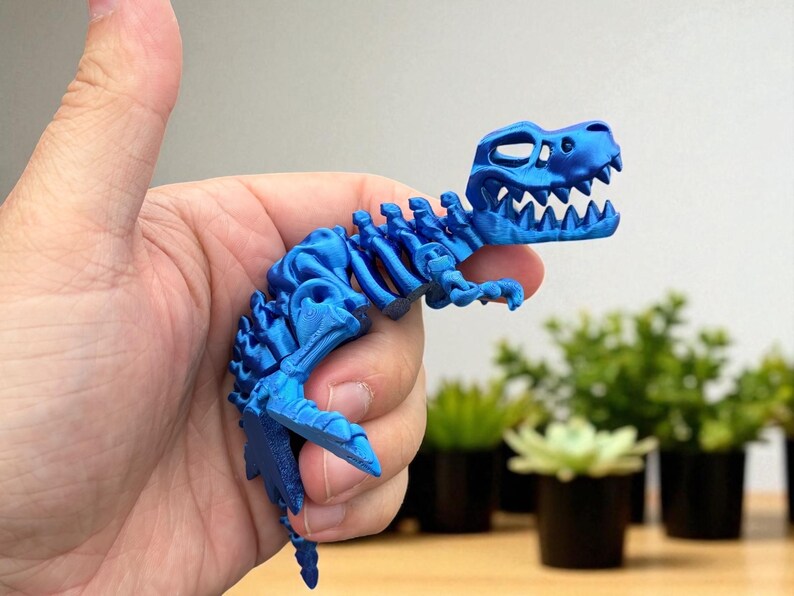 Puede incluir: Un juguete de esqueleto de dinosaurio azul articulado. El juguete es una representaci&oacute;n detallada de un Tyrannosaurus Rex, con costillas, columna vertebral y cr&aacute;neo visibles. El juguete se sostiene en una mano, con un fondo borroso de plantas.