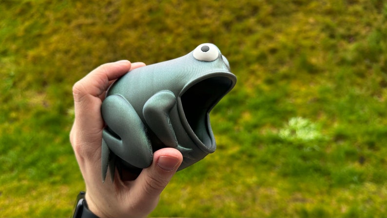 May include: A 3D printed frog figurine with a wide open mouth, painted in a metallic silver and green color. The frog has a white eye with a black pupil.