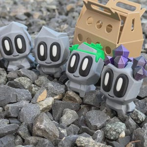 May include: Four gray, geometric figurines with large oval eyes and small smiles, resembling cartoon characters, are displayed on a bed of gray rocks. One figurine has green accents, and another has purple crystal-like structures on its head. A brown cardboard box is in the background.
