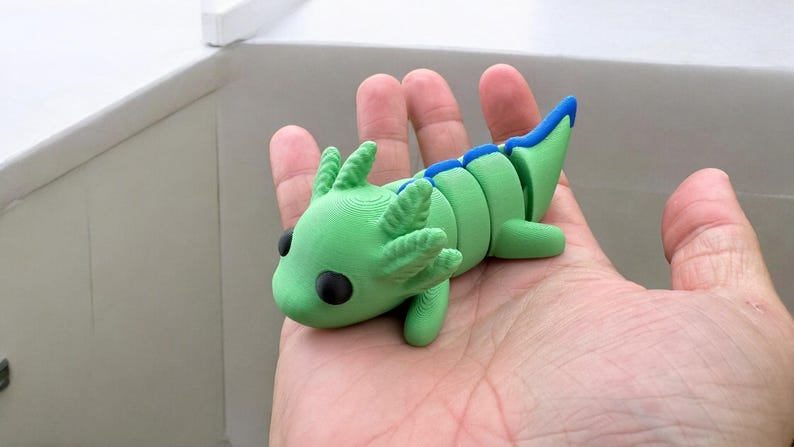 May include: A green and blue 3D printed axolotl figurine. The axolotl has a segmented body and large, black eyes.