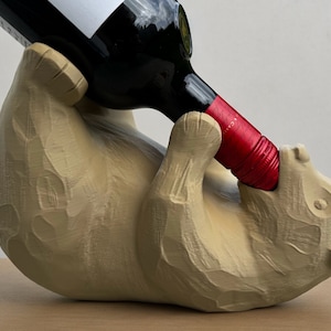 Op de afbeelding: Een houten beren wijnfleshouder met een bruine afwerking. De beer houdt een fles wijn in zijn bek.