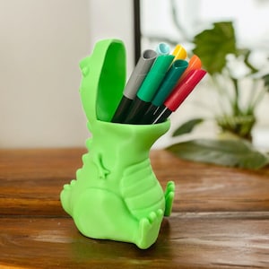 May include: A green dinosaur-shaped pencil holder with a wide open mouth, holding a variety of coloured markers and pens.