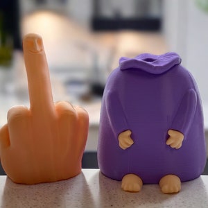 May include: Two 3D printed figurines. One is a purple figure with a rounded body and two small arms. The other is an orange figure shaped like a middle finger with a smiling face.