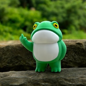 Può includere: Una statuetta di rana verde con occhi gialli e pancia bianca, che fa il dito medio.