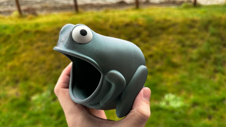 May include: A gray 3D printed frog planter with a large open mouth. The frog has a white eye with a black pupil.