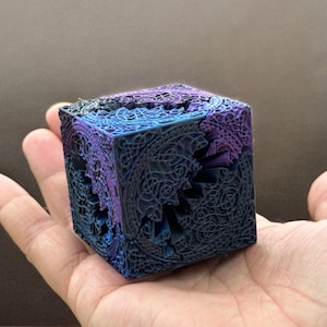 May include: A small, intricately designed cube with a black, blue, and purple color scheme. The cube is made of a 3D printed material and has a complex, geometric pattern.