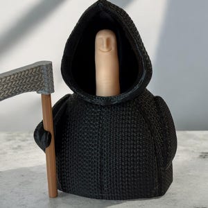 Puede incluir: Una figura negra con capucha y un rostro con forma de dedo sostiene una guadaña plateada con un mango de madera. La figura lleva una túnica negra con capucha y una superficie texturizada.