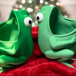 May include: Two green frog-shaped planters with open mouths, facing each other. The planters have large white eyes and are sitting on a red fuzzy surface.
