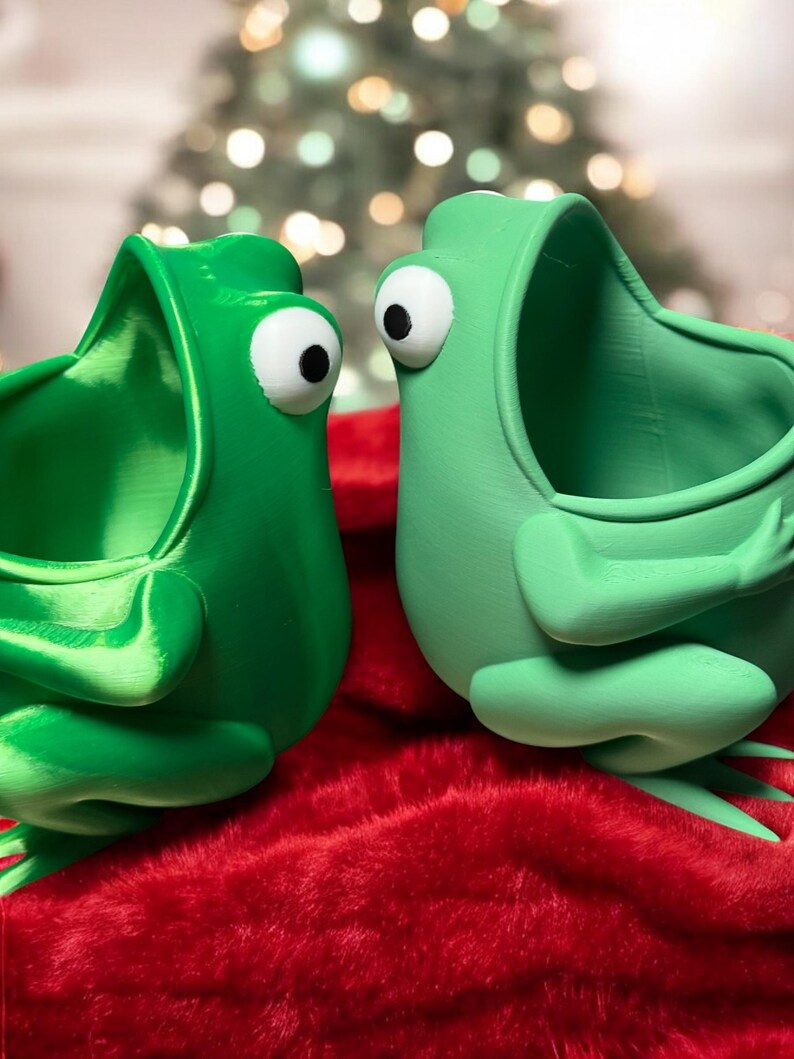 May include: Two green frog-shaped planters with open mouths, facing each other. The planters have large white eyes and are sitting on a red fuzzy surface.