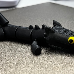 May include: A black 3D printed dragon figurine with yellow eyes. The dragon is posed with its head tilted to the side and its tail curled up.