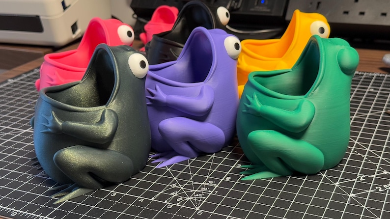 May include: A set of six frog-shaped planters in various colors, including green, purple, black, pink, and yellow. Each planter has a wide open mouth and is designed to hold small plants or succulents.