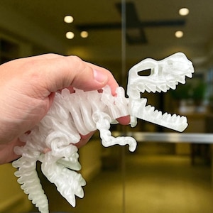 Puede incluir: Un juguete de dinosaurio blanco transl&uacute;cido impreso en 3D, sostenido en una mano. El dinosaurio es un modelo detallado con costillas visibles y una superficie texturizada. El fondo est&aacute; borroso, lo que sugiere un entorno interior.
