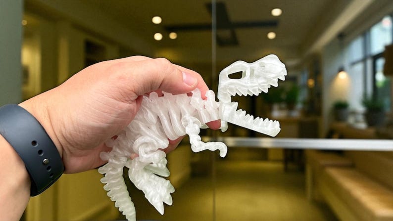 Puede incluir: Un juguete de dinosaurio blanco transl&uacute;cido impreso en 3D, sostenido en una mano. El dinosaurio es un modelo detallado con costillas visibles y una superficie texturizada. El fondo est&aacute; borroso, lo que sugiere un entorno interior.