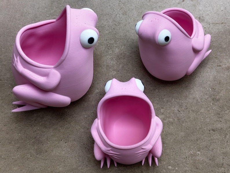 May include: Three pink frog-shaped planters with open mouths. The planters are made of a smooth, matte material and have large, white eyes.