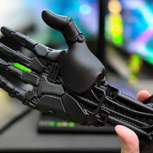 May include: A black 3D printed robotic hand with articulated fingers and a detailed mechanical arm. The hand is open and the arm is extended.