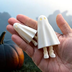 May include: Two white ghost figurines with black eyes, one larger than the other, are held in a hand against a blurry background of pumpkins and fog.