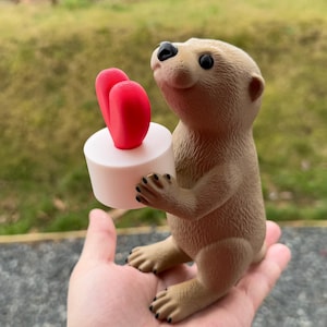May include: A small brown figurine of a meerkat holding a white cake with two pink hearts on top.