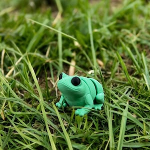 Puede incluir: Una pequeña figura de rana verde sentada en un lecho de hierba verde.