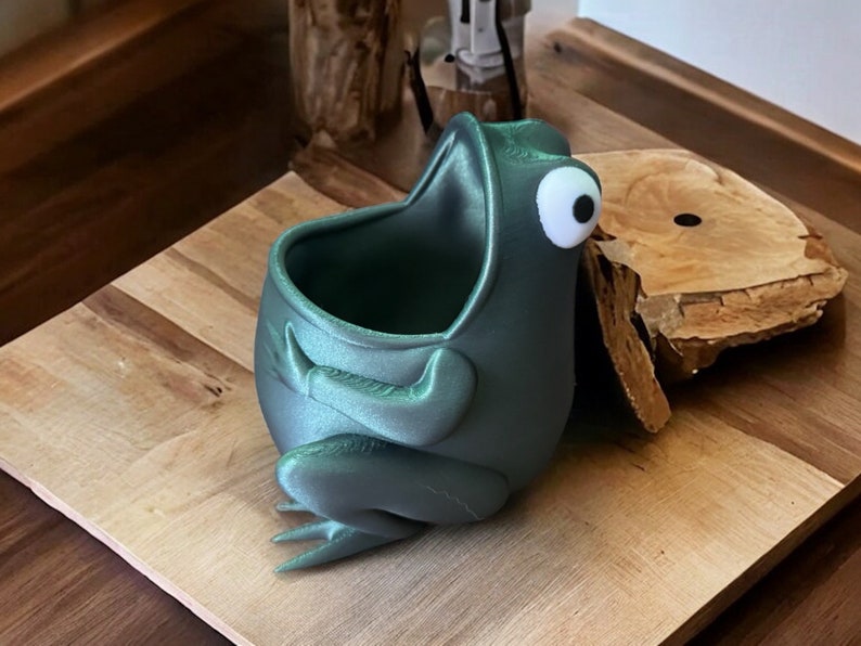 May include: A teal green frog-shaped planter with a large, open mouth and a single white eye. The planter is made of a smooth, plastic-like material and is sitting on a wooden surface.