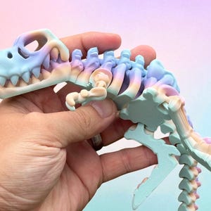 Puede incluir: Un juguete de esqueleto de dinosaurio en colores pastel. El juguete presenta un cr&aacute;neo azul claro con dientes blancos, que transiciona a rosa, morado y amarillo a lo largo de la columna vertebral y la cola. El juguete se sostiene en una mano.
