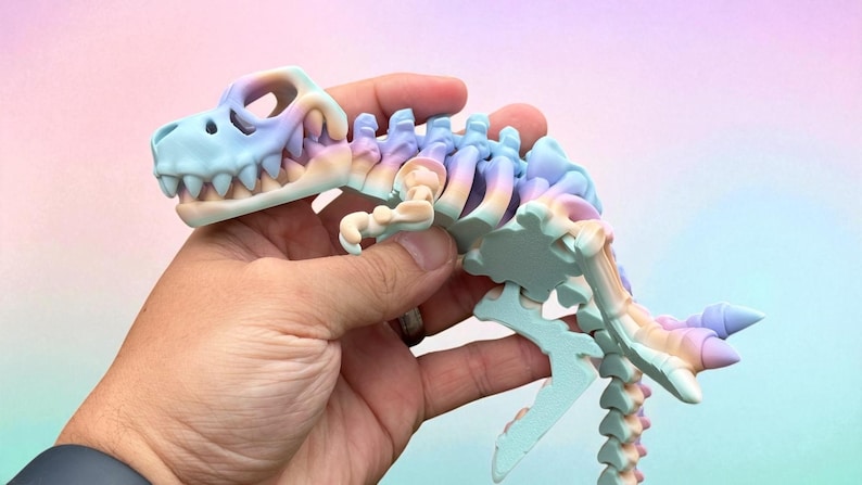 Puede incluir: Un juguete de esqueleto de dinosaurio en colores pastel. El juguete presenta un cr&aacute;neo azul claro con dientes blancos, que transiciona a rosa, morado y amarillo a lo largo de la columna vertebral y la cola. El juguete se sostiene en una mano.