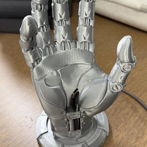 Op de afbeelding: Een zilveren 3D-geprinte robot hand telefoonstandaard met een oplaadkabelgleuf. De hand is geposeerd met de palm naar boven en gespreide vingers. De standaard heeft een ronde basis.
