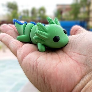 May include: A green and blue axolotl figurine with black eyes. The figurine is sitting in the palm of a hand.