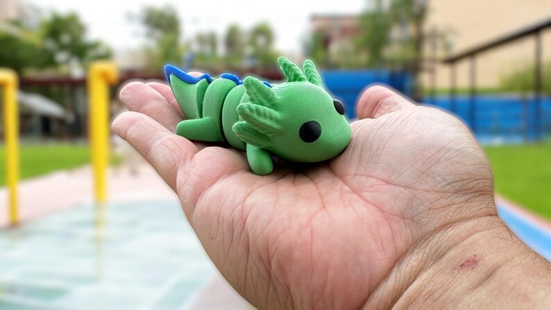 May include: A green and blue axolotl figurine with black eyes. The figurine is sitting in the palm of a hand.