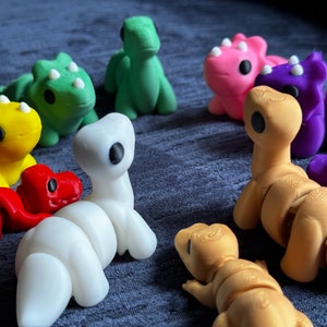 Puede incluir: Una colección de juguetes de dinosaurios impresos en 3D de varios colores. Los dinosaurios están en diferentes poses y colores, incluyendo verde, amarillo, rojo, blanco, rosa, morado y dorado.