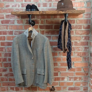 May include: A wooden shelf with black metal pipe brackets mounted on a brick wall. A brown hat, a blazer, and a scarf are hanging on the shelf.