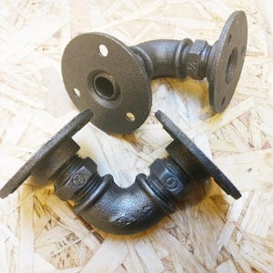 Könnte beinhalten: Zwei schwarze Metall-Regalhalterungen mit Flanschen. Die Halterungen haben die Form eines L und einen Gewindeanschluss.