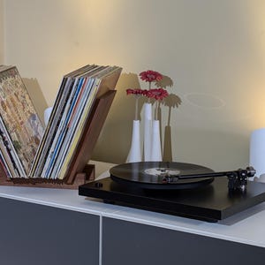 May include: A black turntable with a record on the platter, a wooden record stand with vinyl records, and a white lamp on a white surface.