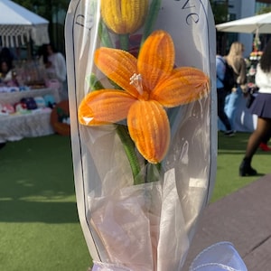 Puede incluir: Un ramo de flores de fieltro amarillo y naranja envuelto en papel de seda blanco y atado con una cinta blanca. Las flores están dispuestas en una bolsa de plástico transparente. La bolsa tiene una etiqueta blanca con el texto "Peace Love" impreso en ella.