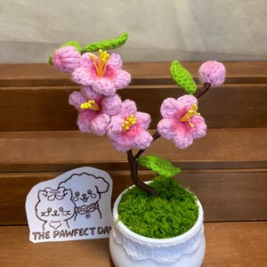 May include: A small crocheted pink cherry blossom tree in a white pot with green moss. The pot has a white lace trim. A sticker with a cartoon dog and cat and the text "The Pawfect Day" is in the foreground.