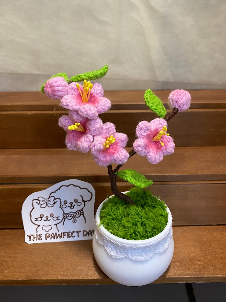 May include: A small crocheted pink cherry blossom tree in a white pot with green moss. The pot has a white lace trim. A sticker with a cartoon dog and cat and the text "The Pawfect Day" is in the foreground.