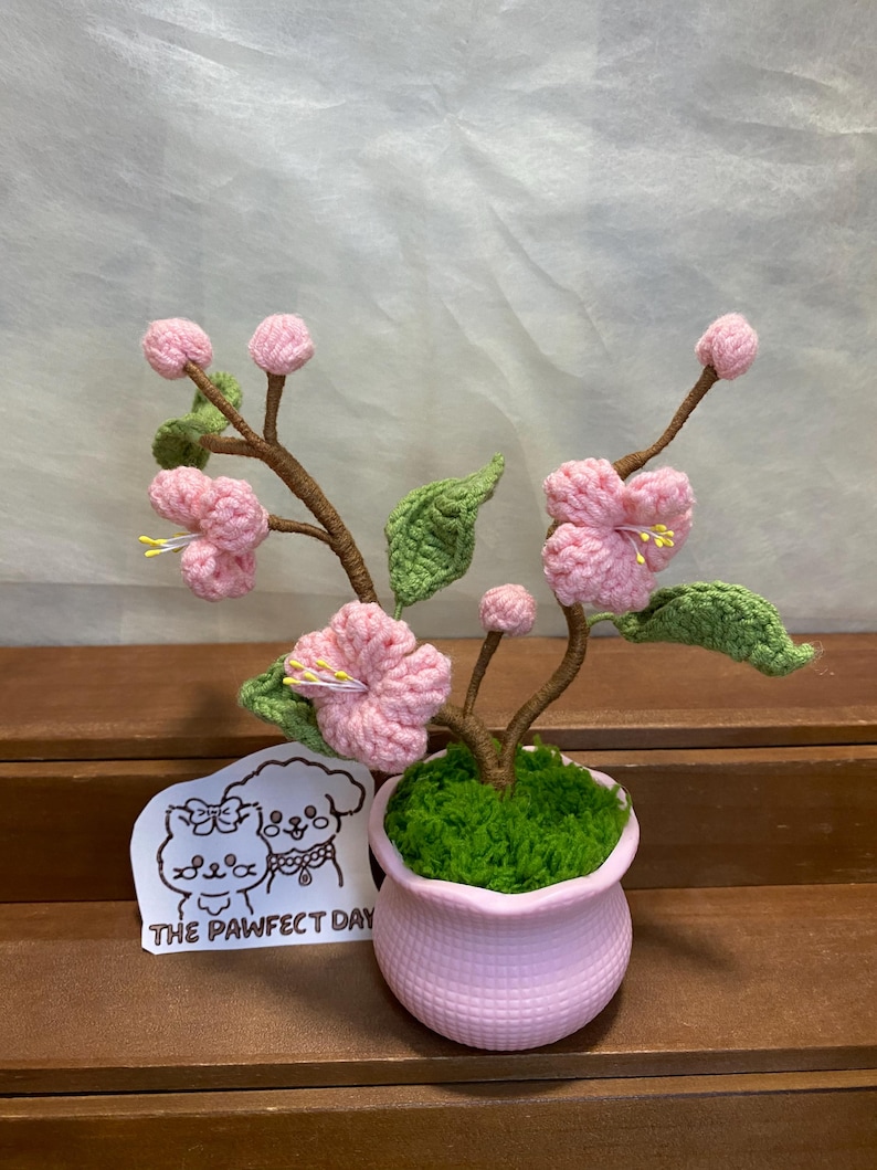 May include: A pink crocheted cherry blossom tree in a pink pot with green moss. The tree is on a wooden surface and has a sticker with a cartoon dog and cat that says "The Pawfect Day".