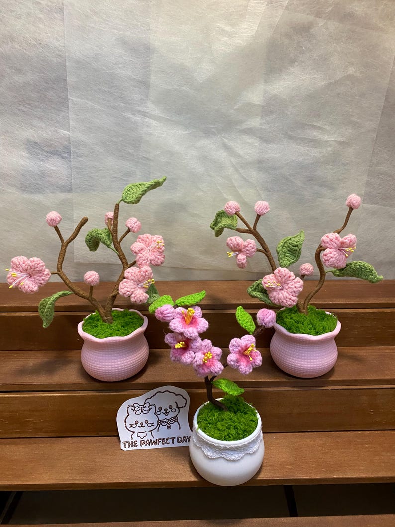 May include: Three crocheted pink cherry blossom trees in pots. The trees are on a wooden shelf. The middle tree is in a white pot with a lace trim. The other two trees are in pink pots. The text "THE PAWFECT DAY" is on a sticker below the middle tree.