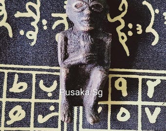 May include: A dark brown, hand-carved figurine of a human figure with a large head and a simple, stylized body. The figurine is sitting on a patterned fabric background with the text "Pusaka.Sg" visible.