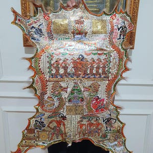 May include: A detailed, hand-painted artwork on a tanned hide, featuring intricate figures, dragons, and ornate patterns. The piece has a scalloped edge with fur trim and is held up against a wall. The artwork is framed in gold.