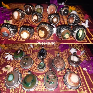 May include: A collection of silver rings and pendants featuring various agate stones. The rings and pendants have intricate designs and feature a variety of colors, including brown, green, white, and red. The stones are set in silver and have a polished finish. The image also includes the text "Pusaka.sg".