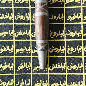 May include: A wooden pendant with silver-colored accents and a black cord. The pendant has a cylindrical shape with a dark brown center and silver bands. The background is a black and yellow patterned fabric with Arabic script.