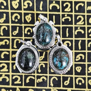 May include: Three silver pendants with oval, dark green and blue gemstones. The pendants have a decorative silver border. The background is a black fabric with a yellow grid pattern and symbols.