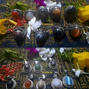 May include: Collection of gemstone rings and pendants. Rings feature various oval-shaped stones in shades of red, brown, gray, and orange, set in silver-toned metal. Pendants with similar stones are also displayed, with the text "Pusaka.sg".