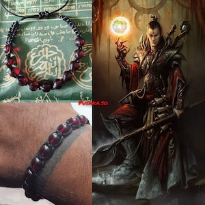 May include: A black beaded bracelet with red glass beads. The bracelet is on a person's wrist. The background is a green fabric with Arabic writing.