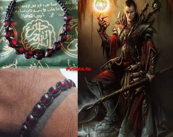 Op de afbeelding: Een zwarte kralen armband met rode glazen kralen. De armband zit om de pols van een persoon. De achtergrond is een groene stof met Arabische tekst.